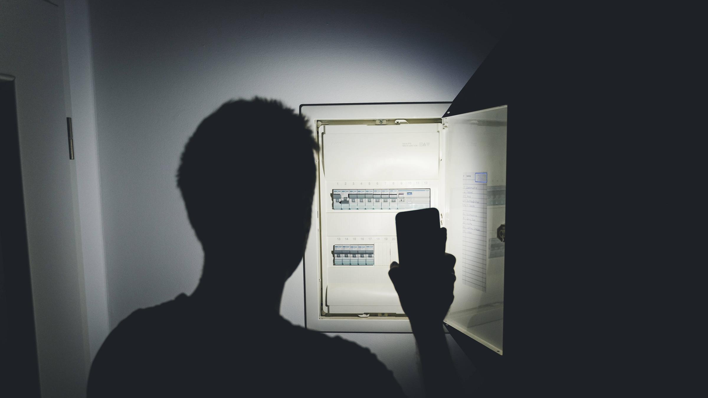 Symboldbild: Stromausfall, ein Mann leuchtet mit seinem Smartphone und prüft den Sicherungskasten
