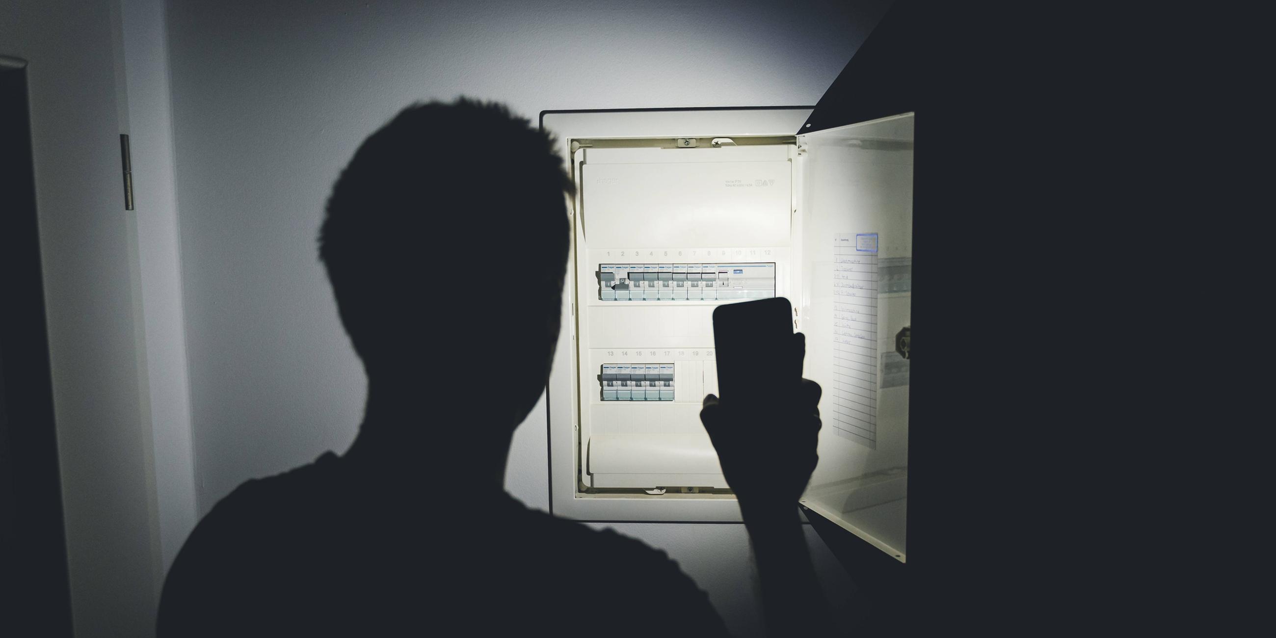 Symboldbild: Stromausfall, ein Mann leuchtet mit seinem Smartphone und prüft den Sicherungskasten