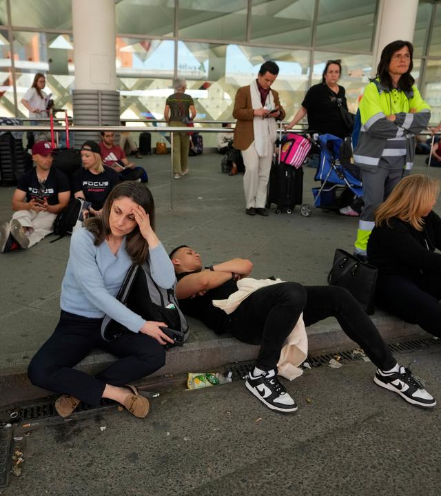 Menschen sitzen und stehen auf einem Bordstein vor dem Bahnhof Madrid-Atocha und warten.