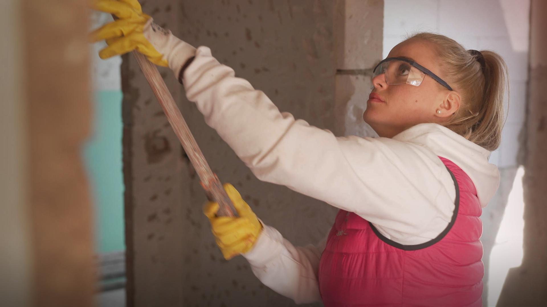 Eine Frau in einer pinkfarbenen Weste arbeitet mit einem Stab an der Wand auf einer Baustelle. 