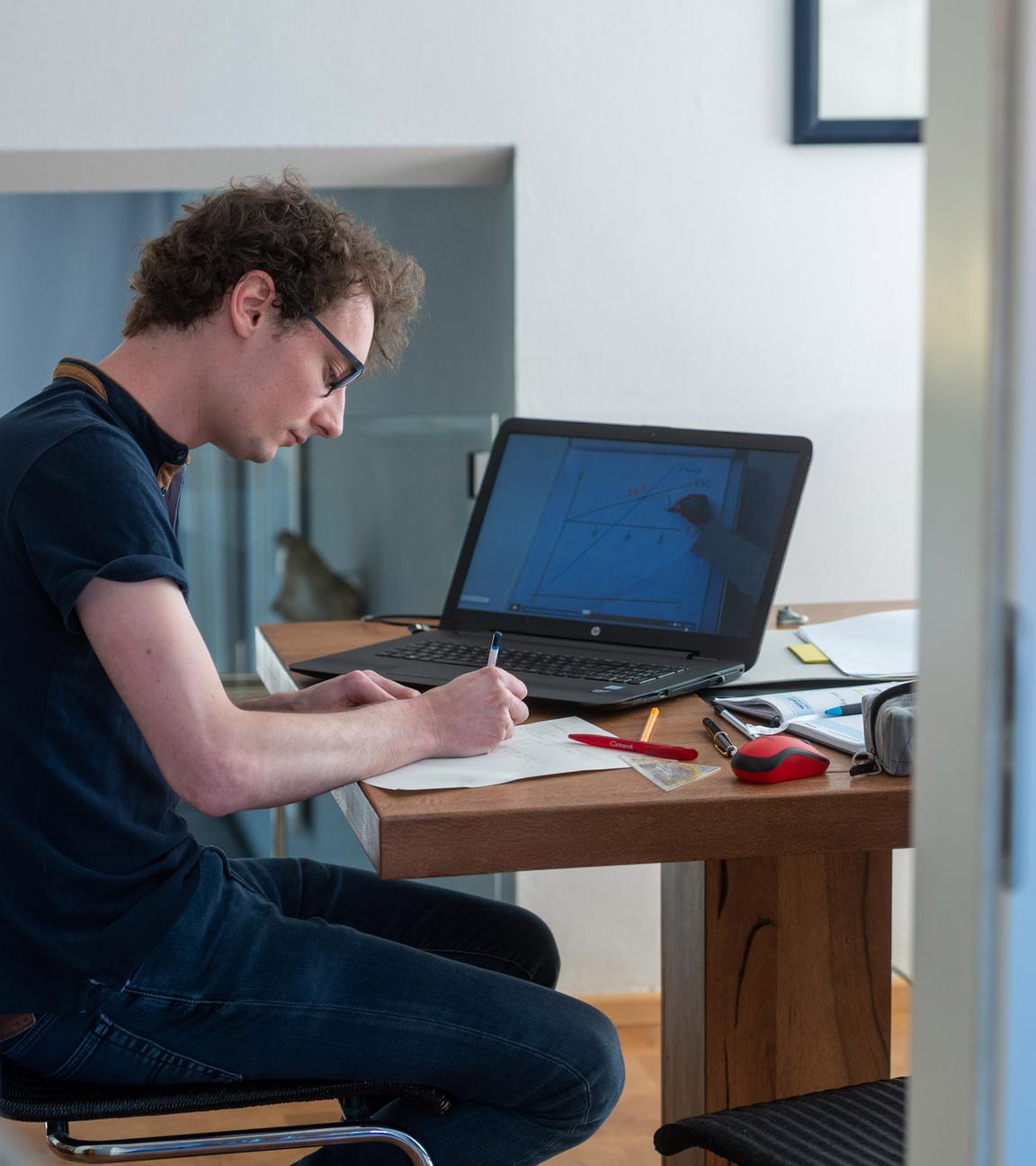 : Ein Student arbeitet zu Hause an seinem Laptop für die Uni. Er schreibt auf einen Zettel, auf dem Laptop läuft eine Vorlesung und um ihn herum auf dem Tisch liegen Stifte, ein Geodreieck, eine Maus, ein Buch und ein Federmäppchen.