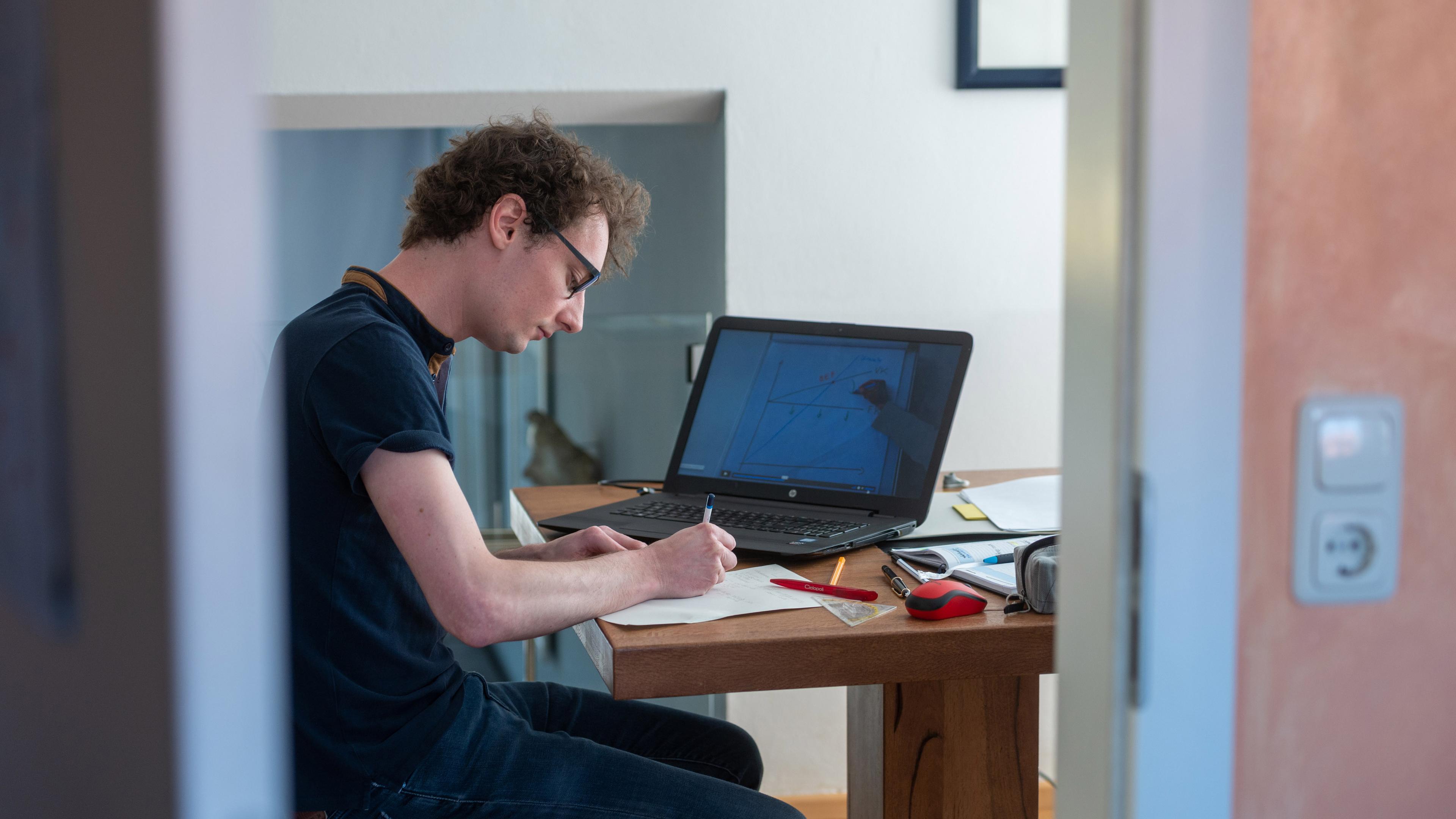 : Ein Student arbeitet zu Hause an seinem Laptop für die Uni. Er schreibt auf einen Zettel, auf dem Laptop läuft eine Vorlesung und um ihn herum auf dem Tisch liegen Stifte, ein Geodreieck, eine Maus, ein Buch und ein Federmäppchen.
