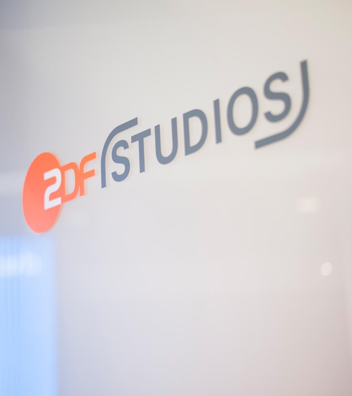 Abgebildet ist das Logo „ZDF Studios“ auf einer glatten Glaswand. Der orangefarbene Kreis mit „ZDF“ ist links platziert, daneben steht „STUDIOS“ in grauer Schrift. Die Umgebung wirkt modern und lichtdurchflutet, Details im Hintergrund sind unscharf