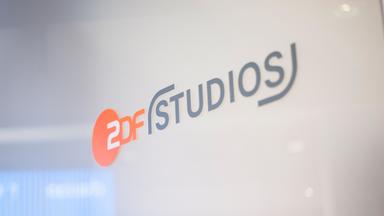 Abgebildet ist das Logo „ZDF Studios“ auf einer glatten Glaswand. Der orangefarbene Kreis mit „ZDF“ ist links platziert, daneben steht „STUDIOS“ in grauer Schrift. Die Umgebung wirkt modern und lichtdurchflutet, Details im Hintergrund sind unscharf