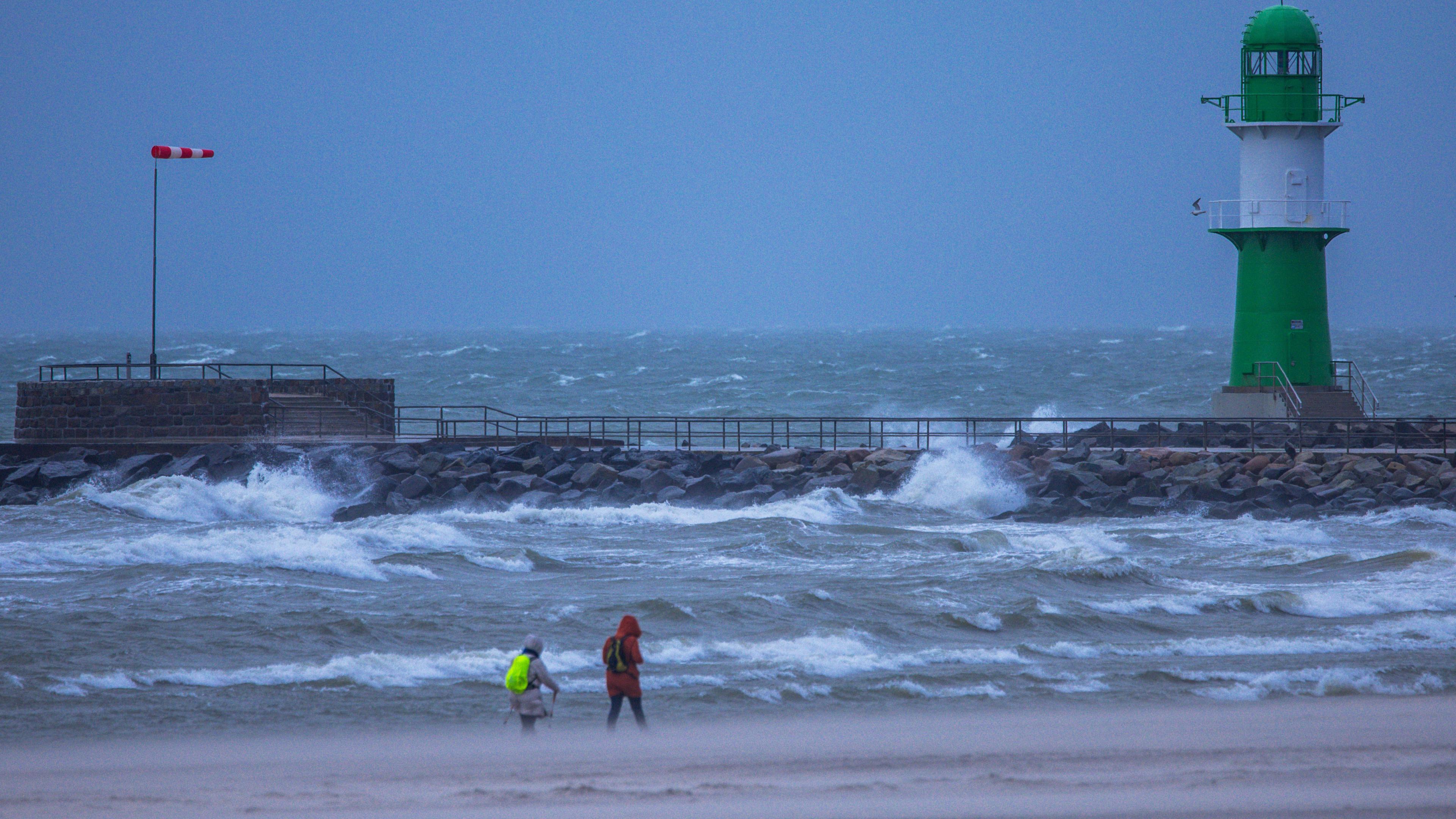 Mecklenburg-Vorpommern, Rostock: Bei stürmischem Wetter sind Spaziergänger vor der Mole an der Ostseeküste unterwegs. Der Sturm bläst Sand vom Strand zum Alten Strom. 