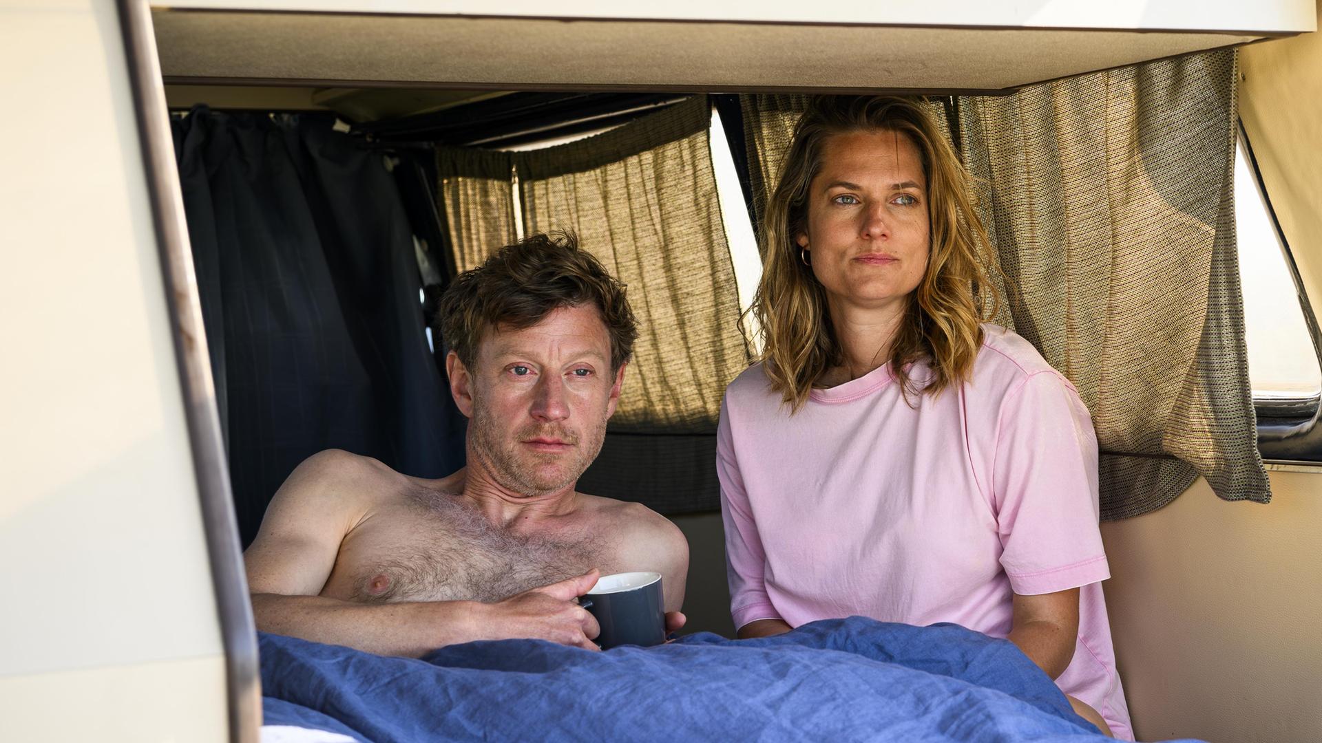 Henk Cassens (Maxim Mehmet) liegt oberkörperfrei unter einer Decke im Van und hält eine Tasse in der Hand. Neben ihm sitzt Anna Mansholt (Marie Burchard) im T-Shirt, beide blicken in die Ferne. Die Heckklappe des Vans ist geöffnet.