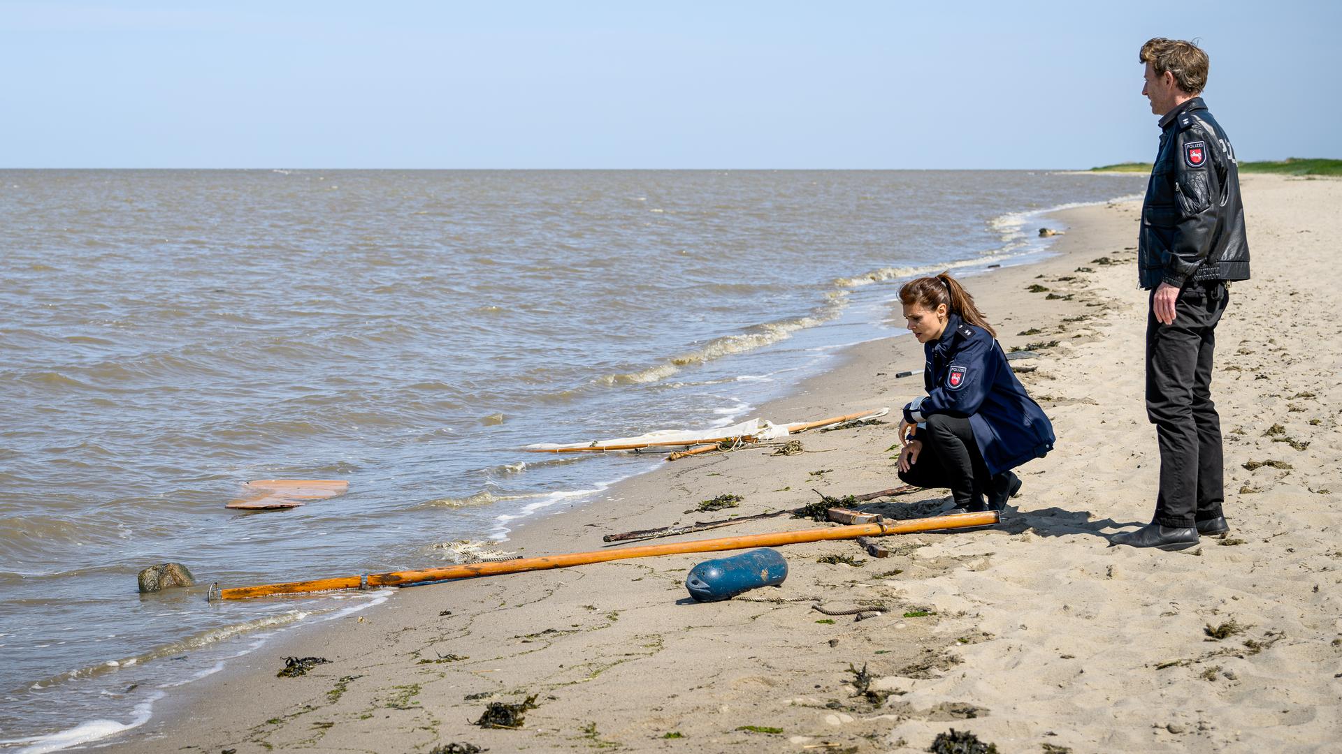 Süher Özlügül (Sophie Dal) kniet am Strand vor zerbrochenen und verstreuten Teilen eines Segelbootes. Hinter ihr steht Henk Cassens (Maxim Mehmet).