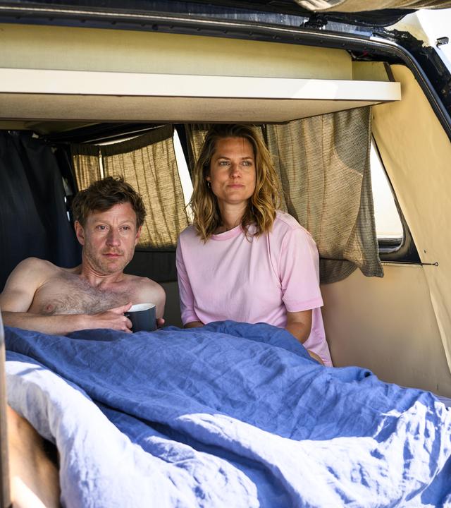 Henk Cassens (Maxim Mehmet) liegt oberkörperfrei unter einer Decke im Van und hält eine Tasse in der Hand. Neben ihm sitzt Anna Mansholt (Marie Burchard) im T-Shirt, beide blicken in die Ferne. Die Heckklappe des Vans ist geöffnet.
