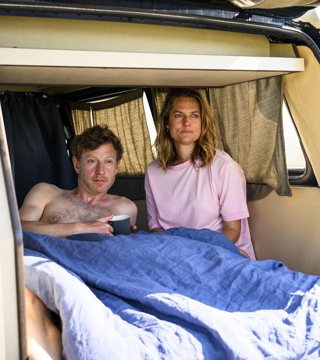 Henk Cassens (Maxim Mehmet) liegt oberkörperfrei unter einer Decke im Van und hält eine Tasse in der Hand. Neben ihm sitzt Anna Mansholt (Marie Burchard) im T-Shirt, beide blicken in die Ferne. Die Heckklappe des Vans ist geöffnet.