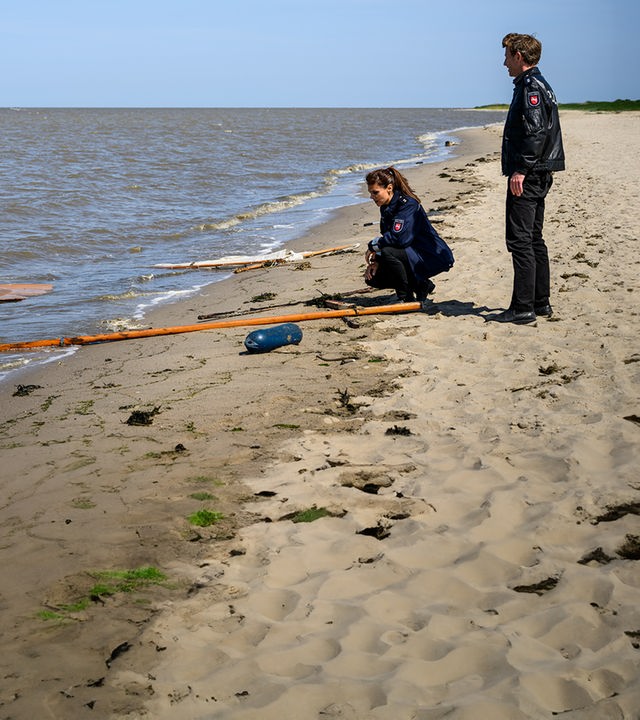 Süher Özlügül (Sophie Dal) kniet am Strand vor zerbrochenen und verstreuten Teilen eines Segelbootes. Hinter ihr steht Henk Cassens (Maxim Mehmet).