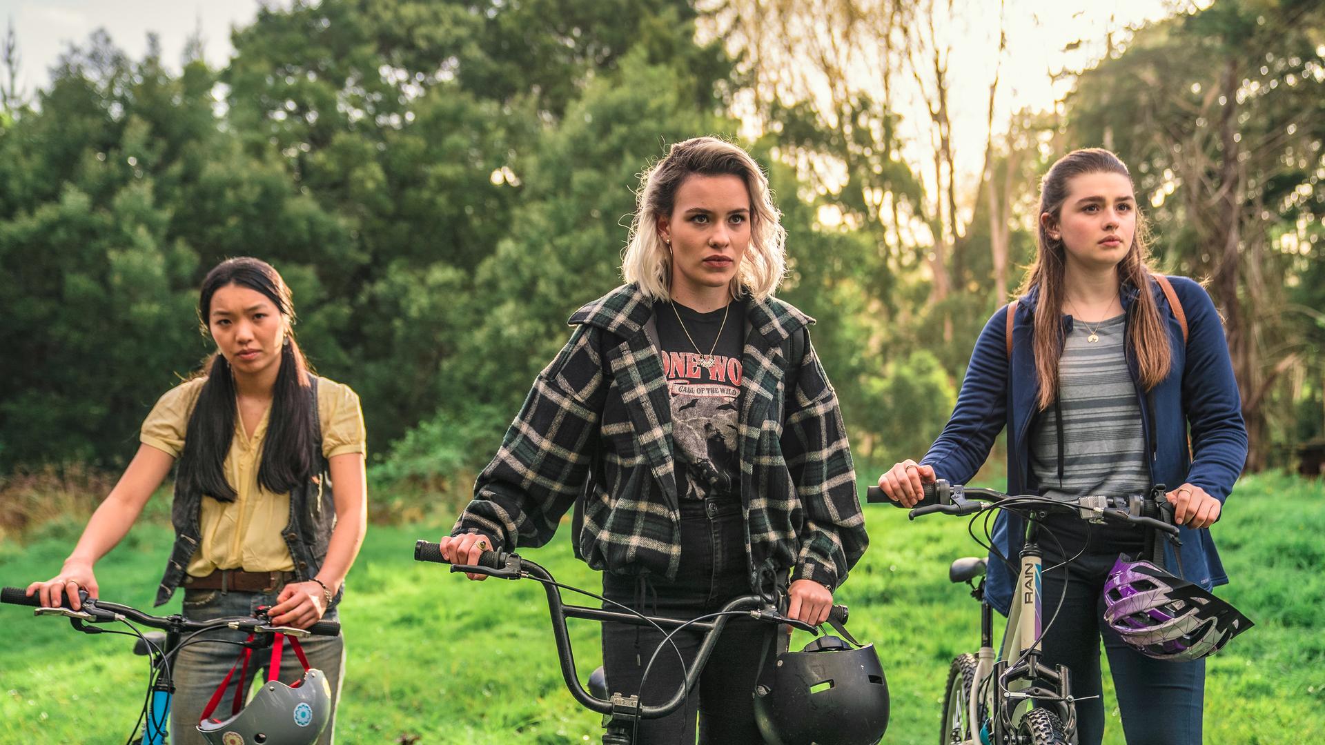 Caroline, Issie und Natasha stehen mit ihren Fahrrädern auf einer Wiese. An den Fahrradlenkern hängen ihre Helme.
