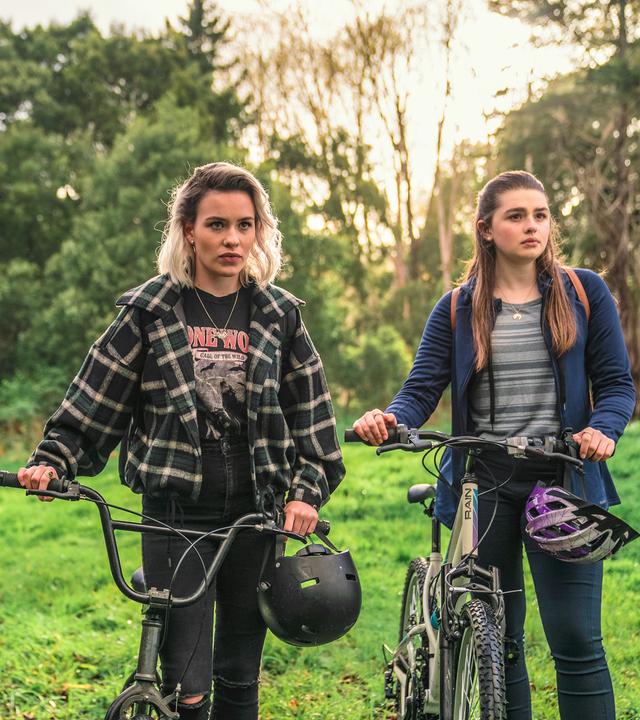 Caroline, Issie und Natasha stehen mit ihren Fahrrädern auf einer Wiese. An den Fahrradlenkern hängen ihre Helme.