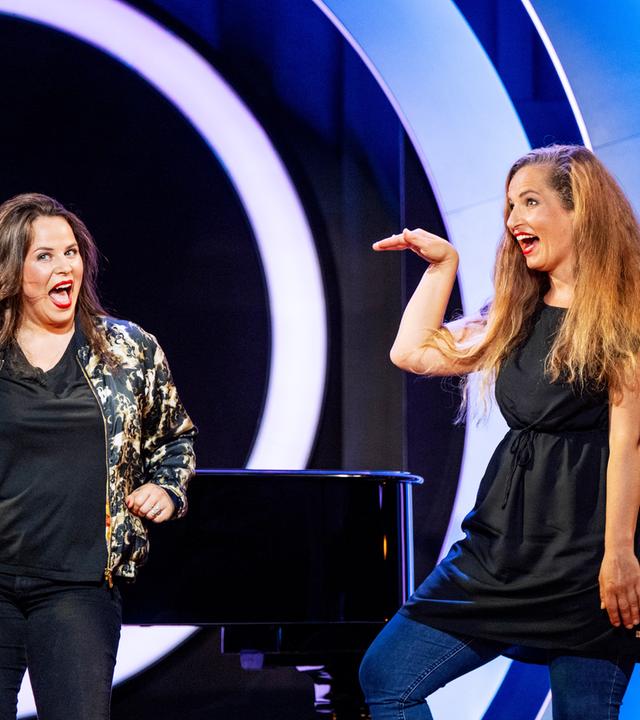 Julia Gamez Martin steht auf der linken Bildseite. Auf der rechten Bildseite ist Ariane Müller zu sehen. Beide haben den Mund geöffnet. Ariane Müller hebt ihre rechte Hand. Julia Gamez Martin stemmt ihre linke Hand in die Hüfte.