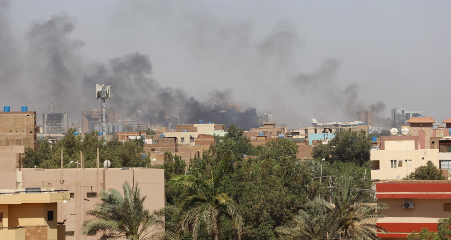 Sudan: Rauch steigt über einer Stadt auf. Im Vordergrund sind Palmen, im Hintergrund Häuser