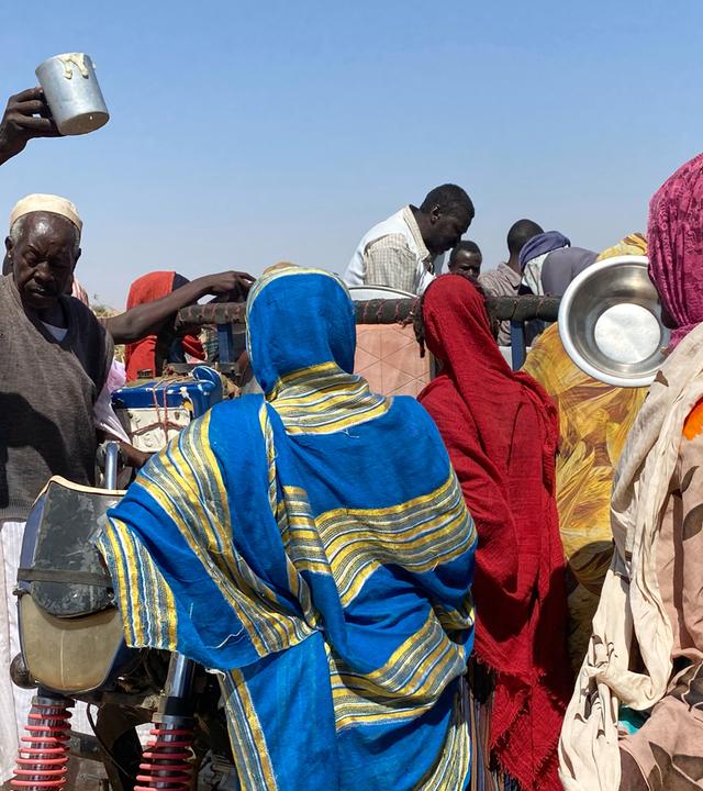 Schwarze Menschen in bunten Gewändern drängen mit leeren Metallschüsseln um einen Wagen, von dem aus Essen verteilt wird.
