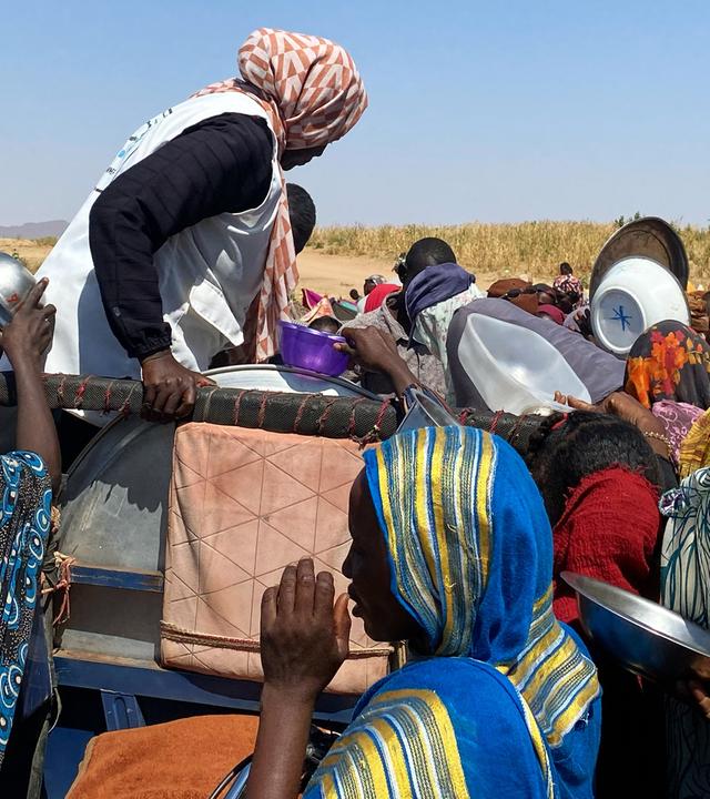 Schwarze Menschen in bunten Gewändern drängen mit leeren Metallschüsseln um einen Wagen, von dem aus Essen verteilt wird.