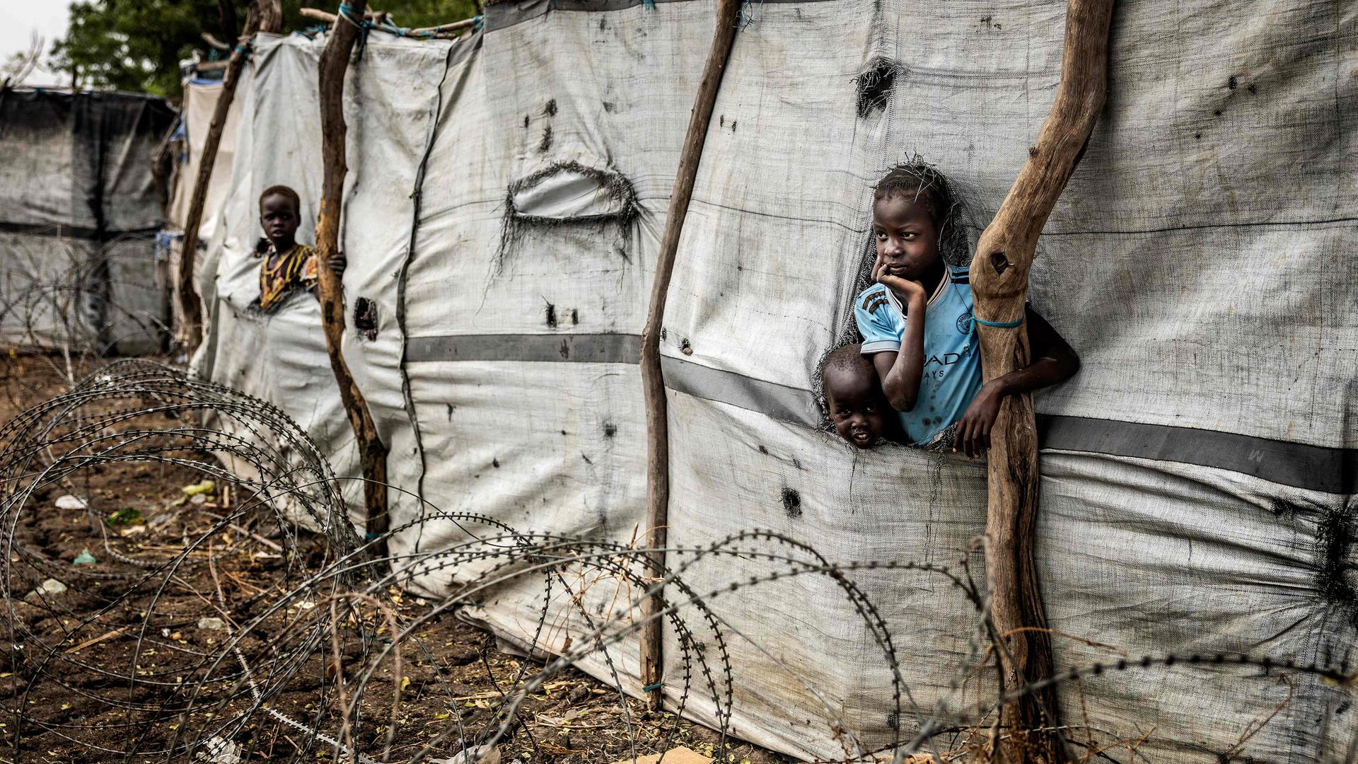 Kinder schauen aus Schlitzen in einer Plane am Zaun eines Flüchtlingslagers.