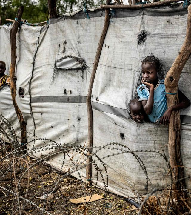Kinder schauen aus Schlitzen in einer Plane am Zaun eines Flüchtlingslagers.