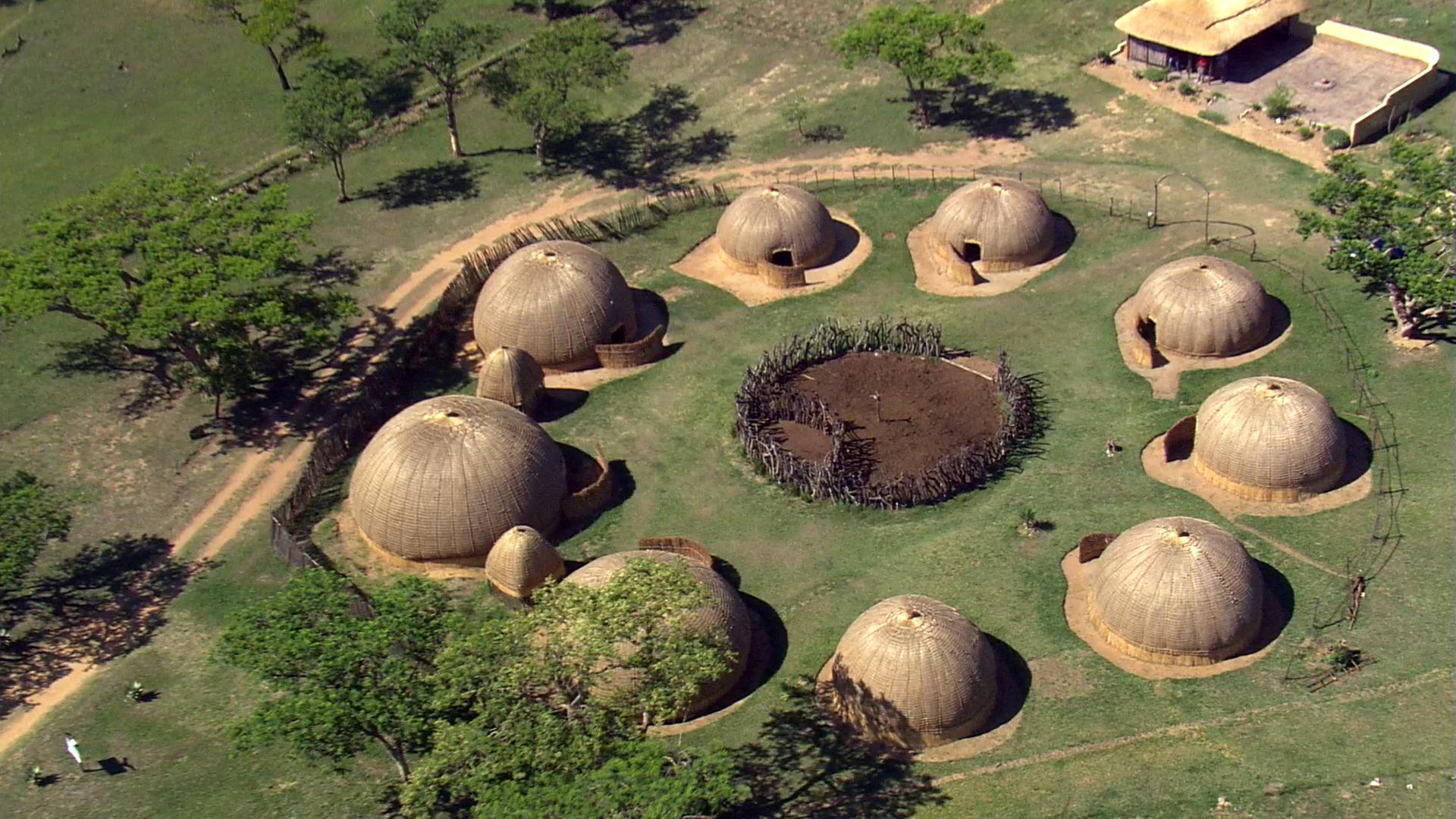 In einem Dorf aus runden Hütten, die im Kreis angeordnet sind, liegt in der Mitte eine Einfriedung. Außen läuft ein Weg entlang an dem einzelne Bäume stehen.
