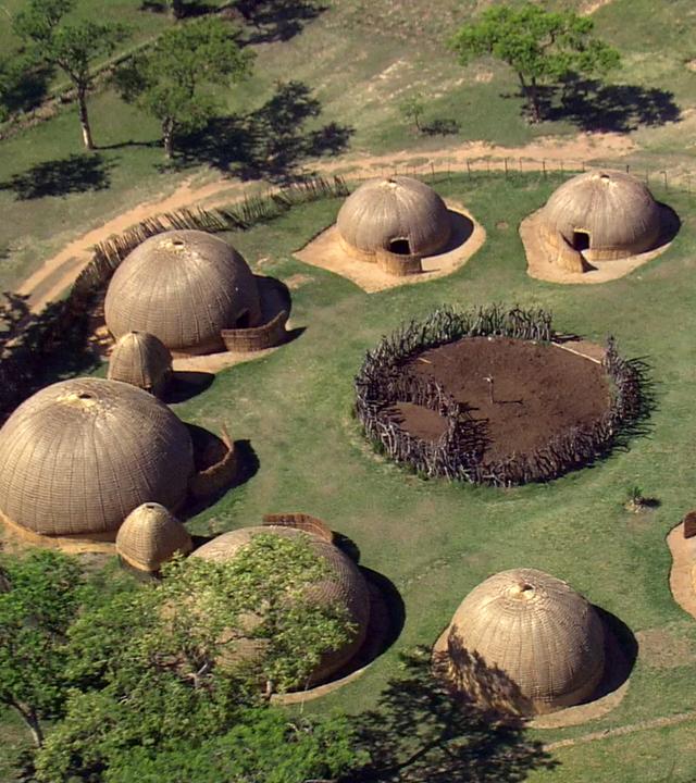 In einem Dorf aus runden Hütten, die im Kreis angeordnet sind, liegt in der Mitte eine Einfriedung. Außen läuft ein Weg entlang an dem einzelne Bäume stehen.