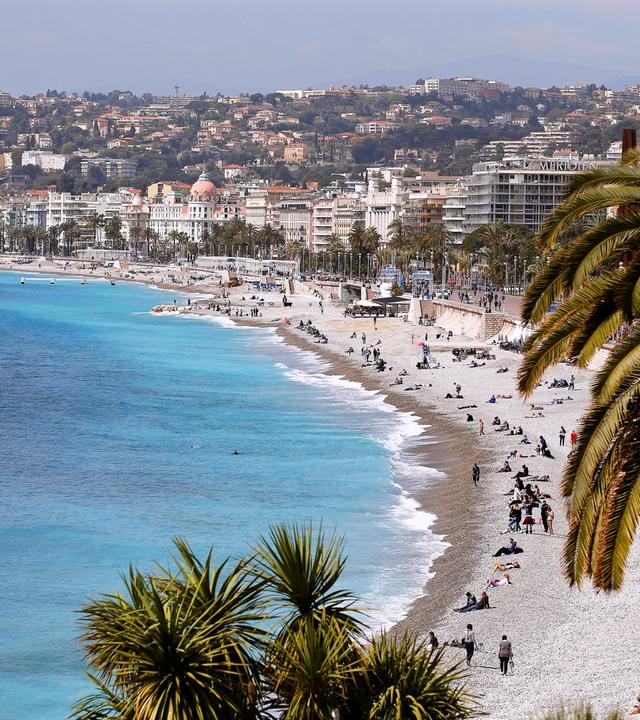 Ansicht der Promenade des Anglais in Nizza, mit Strand und Meer und vielen Häusern.