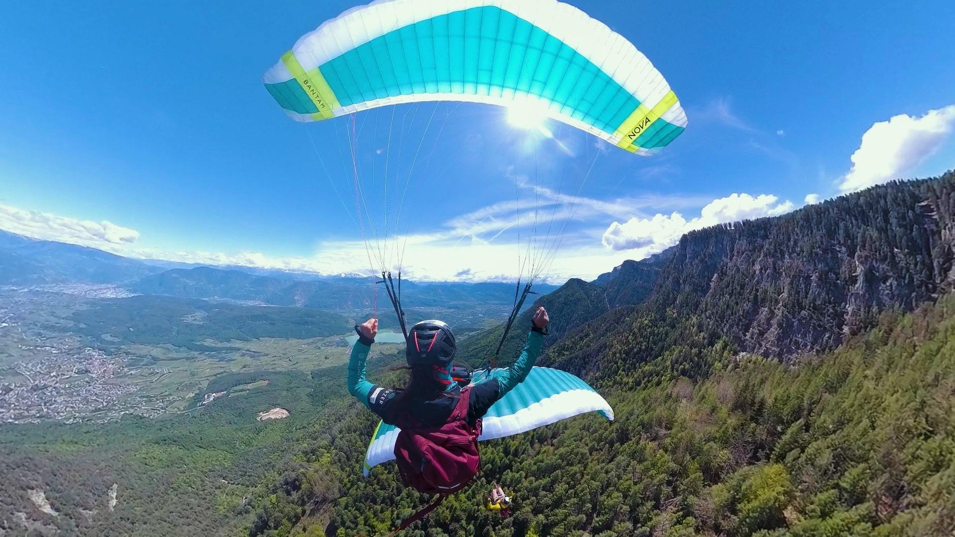 Das Bild zeigt eine Person, die mit einem Gleitschirm in der Luft schwebt. Die Perspektive ist von oben, sodass man die weite Landschaft darunter sehen kann. Im Hintergrund erstrecken sich grüne Hügel und Berge, die von einem dichten Wald bedeckt sind. Der Himmel ist blau mit vereinzelt weißen Wolken, und die Sonne scheint hell. Die Person trägt eine gefärbte Jacke und einen Helm, während die Hände triumphierend in die Höhe gestreckt sind. Der Gleitschirm hat eine zweifarbige Bespannung in Blau und Weiß mit einem gelben Streifen. Unten in der Landschaft sind geordnete Felder und Wohngebiete zu erkennen, die sich entlang der Mitte des Bildes erstrecken. Die gesamte Szene vermittelt ein Gefühl von Freiheit und Abenteuer in einer beeindruckenden Berglandschaft.