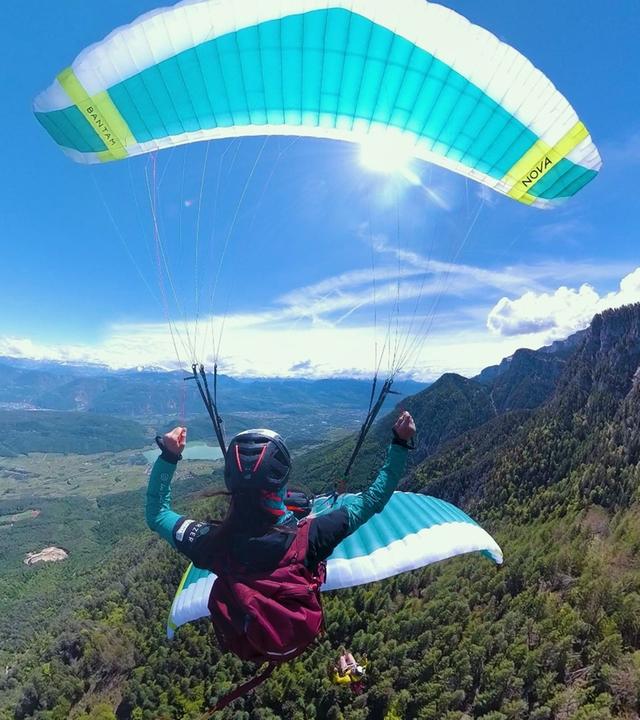 Das Bild zeigt eine Person, die mit einem Gleitschirm in der Luft schwebt. Die Perspektive ist von oben, sodass man die weite Landschaft darunter sehen kann. Im Hintergrund erstrecken sich grüne Hügel und Berge, die von einem dichten Wald bedeckt sind. Der Himmel ist blau mit vereinzelt weißen Wolken, und die Sonne scheint hell. Die Person trägt eine gefärbte Jacke und einen Helm, während die Hände triumphierend in die Höhe gestreckt sind. Der Gleitschirm hat eine zweifarbige Bespannung in Blau und Weiß mit einem gelben Streifen. Unten in der Landschaft sind geordnete Felder und Wohngebiete zu erkennen, die sich entlang der Mitte des Bildes erstrecken. Die gesamte Szene vermittelt ein Gefühl von Freiheit und Abenteuer in einer beeindruckenden Berglandschaft.