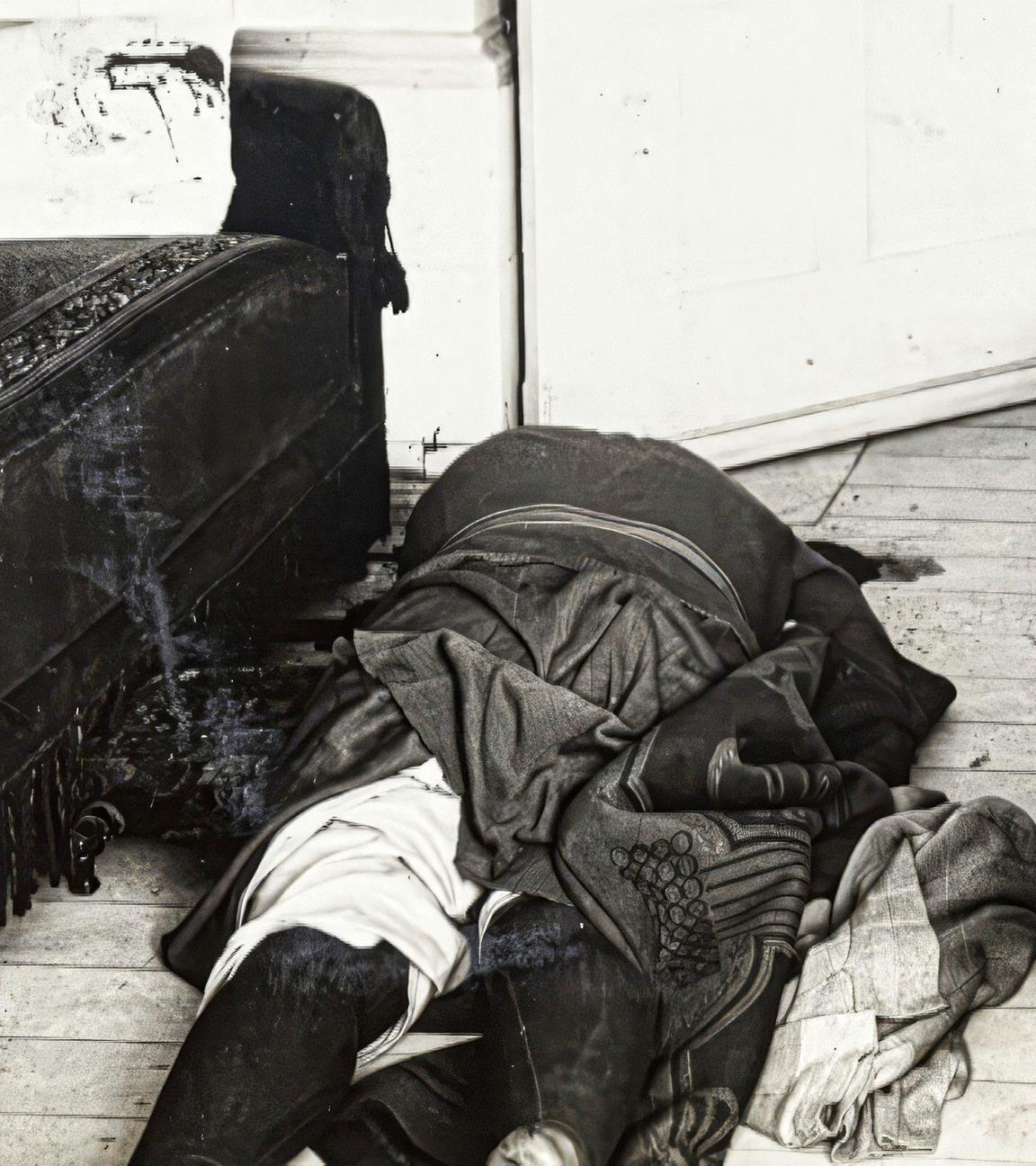 Schwarz-Weiß-Foto: Eine tote Frau liegt auf dem Boden ihres Wohnzimmers. Neben ihr steht auf einer Seite ein Sofa, auf der anderen ein Tisch.