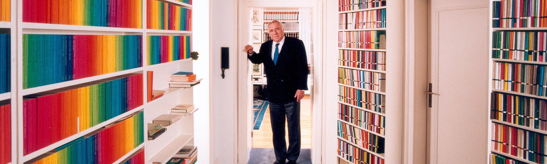 Der Verleger Siegfried Unseld steht vor Bücherregalen. Es sind die Bücher der edition suhrkamp in Regenbogenfarben.