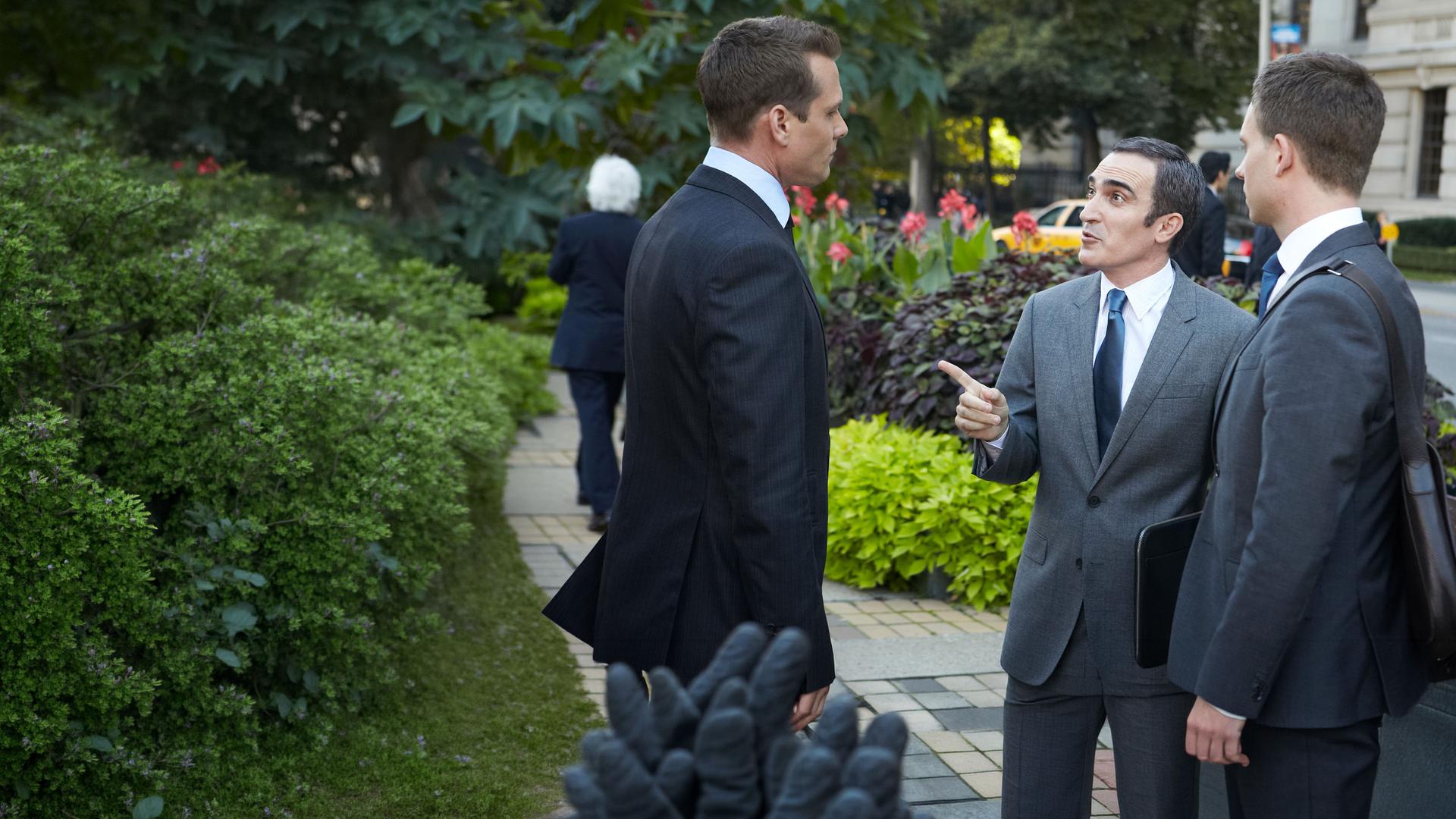 "Suits - Spannungsfelder": Harvey (Gabriel Macht) und Mike (Patrick J. Adams) stehen mit Elliott Stemple (Patrick Fischler) auf dem Gehweg vor einem Gebäude. Elliott Stemple (Patrick Fischler) zeigt gestikulierend auf Harvey. Hinter ihnen ist ein Beet mit Grünpflanzen, am Straßenrand stehen Bäume und Passanten (Komparsen) laufen die Straße mit parkenden Autos entlang.