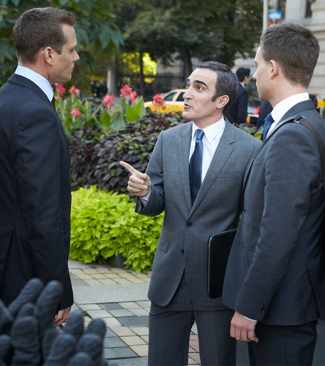 "Suits - Spannungsfelder": Harvey (Gabriel Macht) und Mike (Patrick J. Adams) stehen mit Elliott Stemple (Patrick Fischler) auf dem Gehweg vor einem Gebäude. Elliott Stemple (Patrick Fischler) zeigt gestikulierend auf Harvey. Hinter ihnen ist ein Beet mit Grünpflanzen, am Straßenrand stehen Bäume und Passanten (Komparsen) laufen die Straße mit parkenden Autos entlang.