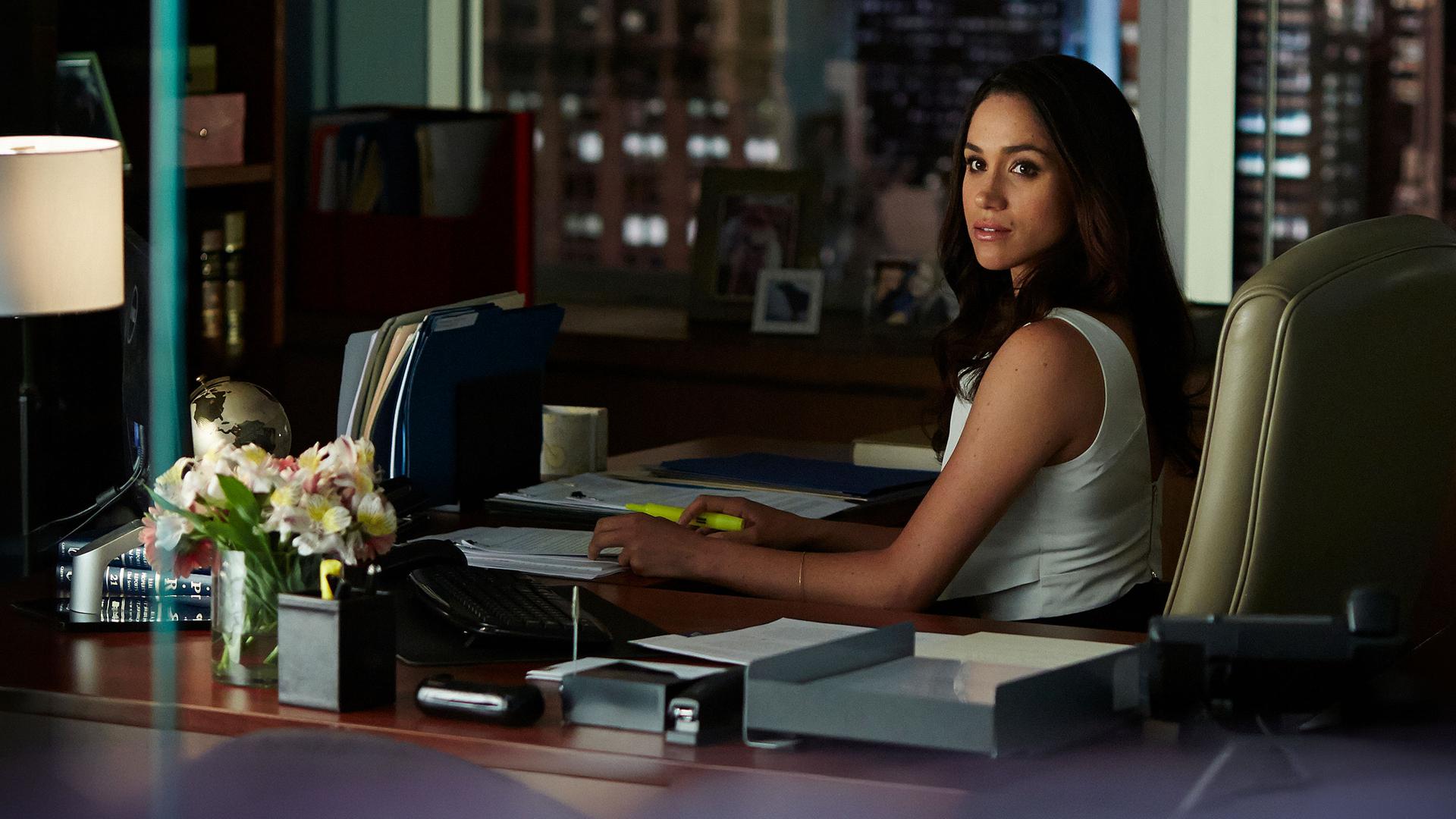 "Suits - Tiefe Einblicke": Rachel (Meghan Markle) sitzt in einem stilvoll eingerichteten Büro am Schreibtisch. Sie hat beide Arme auf der Tischplatte gelegt und schaut erwartungsvoll über ihre linke Schulter.
