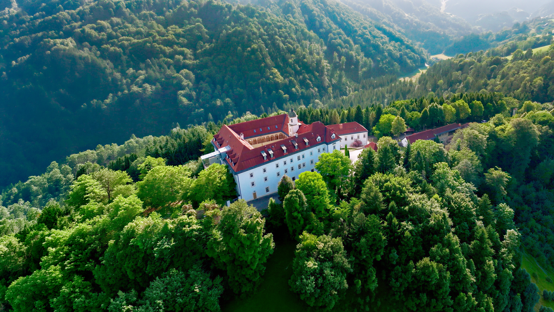 Das Bild zeigt eine malerische Landschaft im Sulmtal, Weststeiermark, Österreich. Im Vordergrund befindet sich ein großes, dreigeschossiges Gebäude mit weißen Wänden und roten Dächern. Das Gebäude ist umgeben von üppigem Grün; Bäume und Sträucher bilden eine dichte Vegetation. In der oberen rechten Bildhälfte sind sanfte Hügel zu sehen, die ebenfalls bewaldet sind.  Hinter dem Gebäude schlängelt sich ein Fluss durch das Tal. Die Landschaft ist von sanften Hügeln und bewaldeten Bereichen geprägt, was ein ruhiges und idyllisches Bild vermittelt. Die Lichtverhältnisse deuten auf einen sonnigen Tag hin, wobei die Sonne durch die Bäume scheint und das Bild aufhellt.   Das Bild könnte aus der ORF/3sat-Dokumentation über das Sulmtal stammen, die die Natur und Kultur der Region thematisiert.