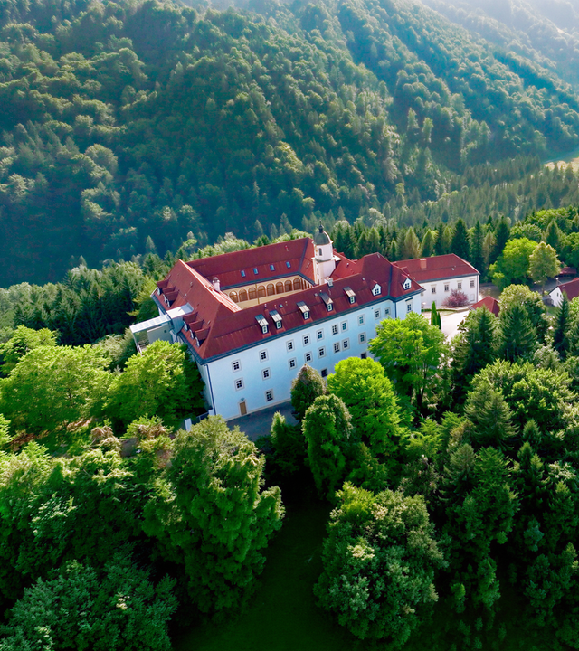 Das Bild zeigt eine malerische Landschaft im Sulmtal, Weststeiermark, Österreich. Im Vordergrund befindet sich ein großes, dreigeschossiges Gebäude mit weißen Wänden und roten Dächern. Das Gebäude ist umgeben von üppigem Grün; Bäume und Sträucher bilden eine dichte Vegetation. In der oberen rechten Bildhälfte sind sanfte Hügel zu sehen, die ebenfalls bewaldet sind.  Hinter dem Gebäude schlängelt sich ein Fluss durch das Tal. Die Landschaft ist von sanften Hügeln und bewaldeten Bereichen geprägt, was ein ruhiges und idyllisches Bild vermittelt. Die Lichtverhältnisse deuten auf einen sonnigen Tag hin, wobei die Sonne durch die Bäume scheint und das Bild aufhellt.   Das Bild könnte aus der ORF/3sat-Dokumentation über das Sulmtal stammen, die die Natur und Kultur der Region thematisiert.