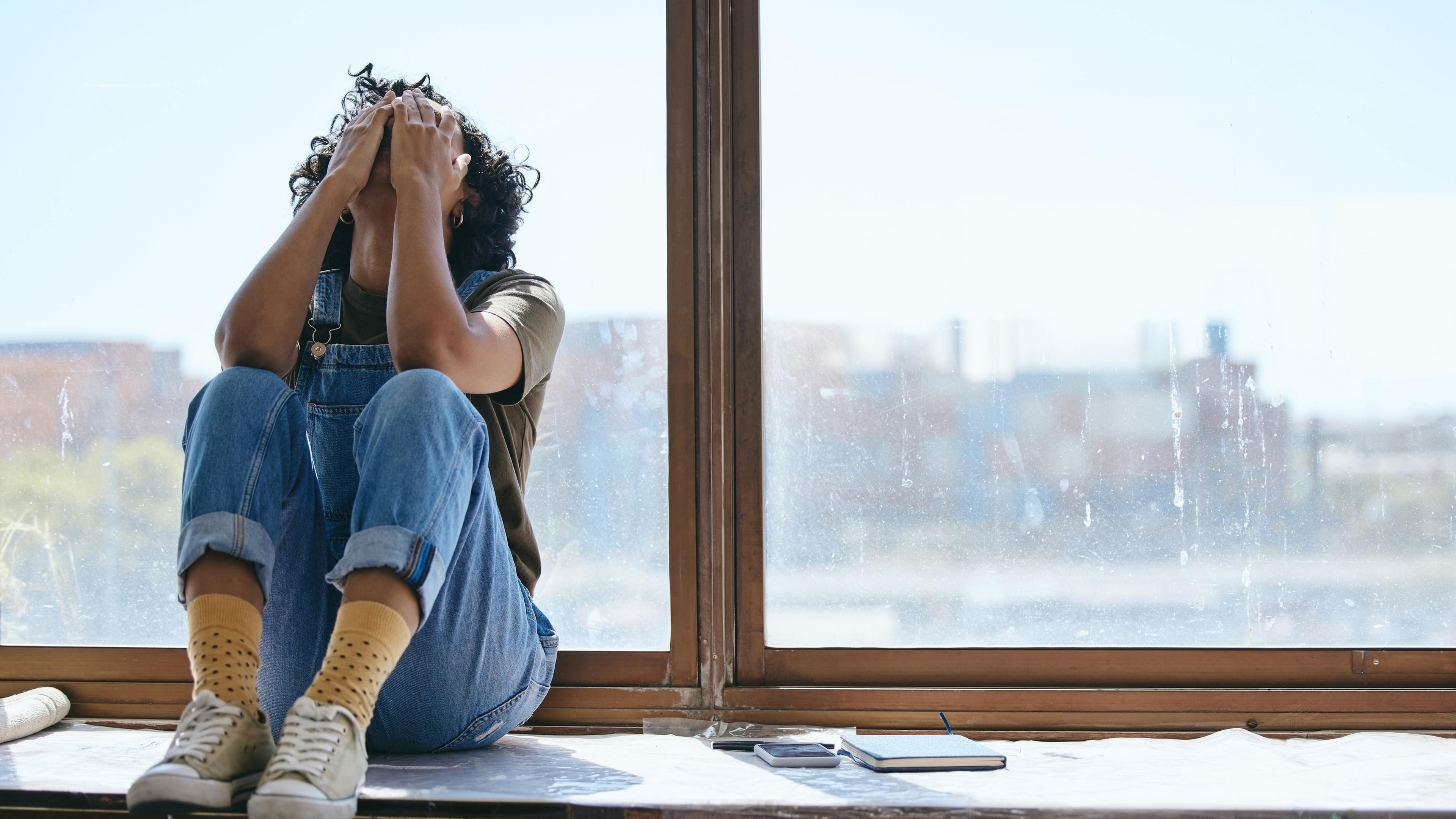 Eine junge Frau sitzt auf einer Fensterbank und legt verzweifelt die Hände in den Kopf