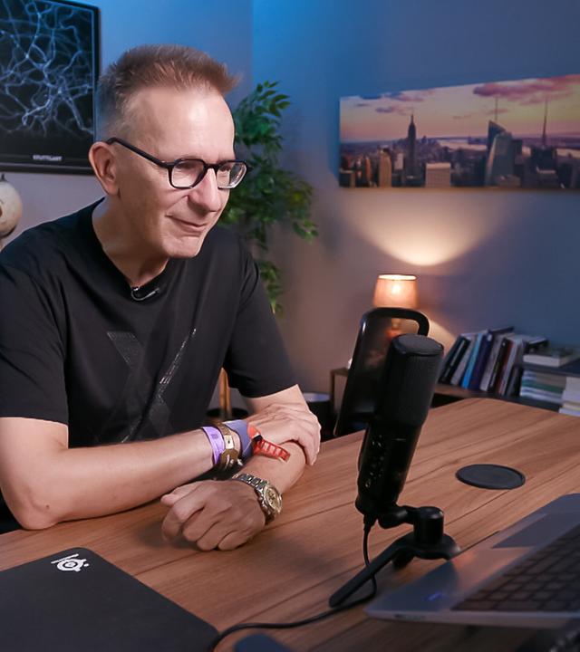 Das Bild zeigt einen Mann, der am Schreibtisch sitzt und an einem Computer arbeitet. Er trägt eine schwarze T-Shirt mit einem dezenten Muster und eine Brille mit dünnem Rahmen. Auf seinem Arm sind mehrere Armbänder sichtbar, und am Handgelenk trägt er eine Armbanduhr.  Der Schreibtisch ist aus Holz und darauf befindet sich ein Mikrofon, ein Laptop und ein schwarzes Mauspad. Im Hintergrund sind Regale mit Büchern und ein Globus zu sehen. An der Wand hängen zwei Poster mit abstrakten Karten von Städten (Frankfurt und Stuttgart). Neben steht eine Lampe, die eine angenehme Beleuchtung bietet, und an der Wand gegenüber ist ein Bild einer Skyline mit Hochhäusern in der Dämmerung.  Die gesamte Szene vermittelt eine moderne und professionelle Atmosphäre, die typisch für einen Arbeitsbereich ist.