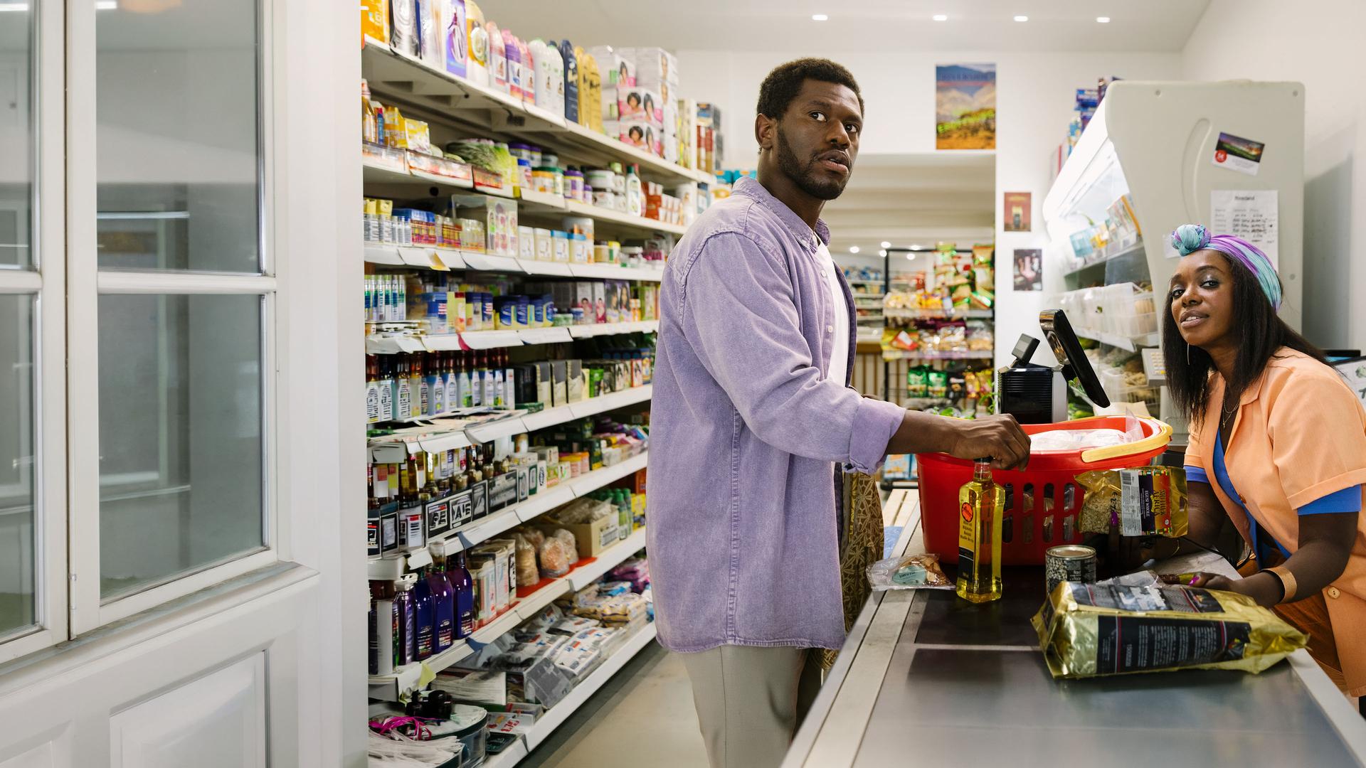 "I don't work here - Supermarkt": Dawit (Akeem van Flodrop) steht an der Kasse in einem Supermarkt. Hinter ihm steht ein gut gefülltes Verkaufsregal. Mit der rechten Hand stellt er eine Flasche auf das vor ihm laufende Kassenband, auf dem auch ein vollgepackter roter Einkaufskorb steht. Skeptisch schaut er zur rechten Seite. In die gleiche Richtung schaut auch die Kassiererin (Karmela Shako), die auf der anderen Seite des Kassenbandes sitzt und sich leicht nach vorne lehnt. Sie trägt einen hellorangenen Kittel und einen Turban, der über ihrer Stirn geknotet ist.