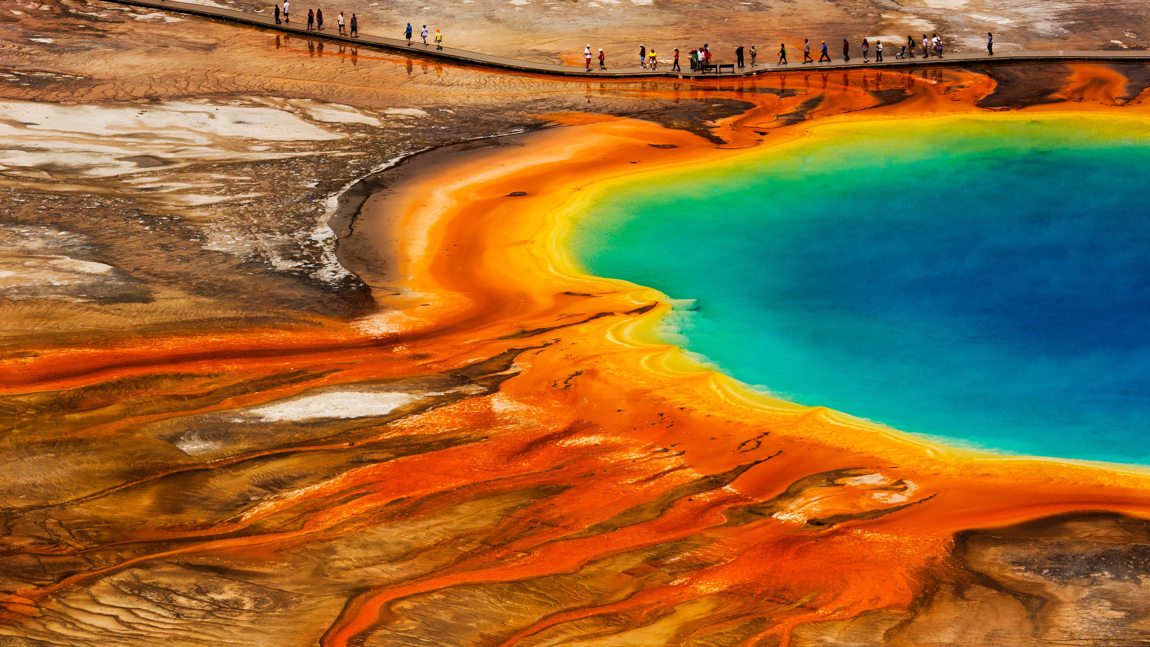 Blick von schräg oben auf den "Grand Prismatic Spring": Ein großer Krater mit orangefarbenem Rand ist gefüllt mit grünem und blauem Wasser. Im Hintergrund laufen Menschen auf einem Steg.