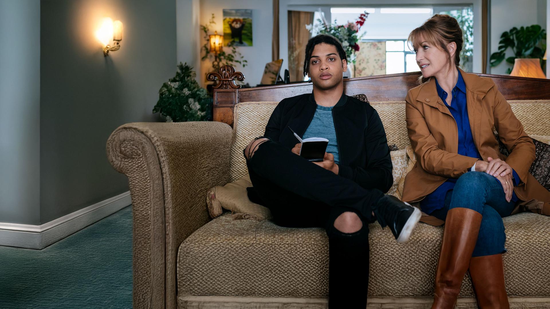 "Harry Wild - Mörderjagd in Dublin: Susans Perücke ... verzweifelt gesucht": Harry Wild (Jane Seymour) sitzt auf einem beigefarbenen Sofa und lächelt den neben ihr sitzenden Fergus (Rohan Nedd) zu. Fergus hält einen Notizblock in seinen Händen.