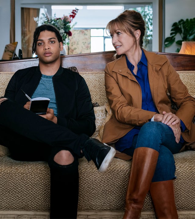 "Harry Wild - Mörderjagd in Dublin: Susans Perücke ... verzweifelt gesucht": Harry Wild (Jane Seymour) sitzt auf einem beigefarbenen Sofa und lächelt den neben ihr sitzenden Fergus (Rohan Nedd) zu. Fergus hält einen Notizblock in seinen Händen.