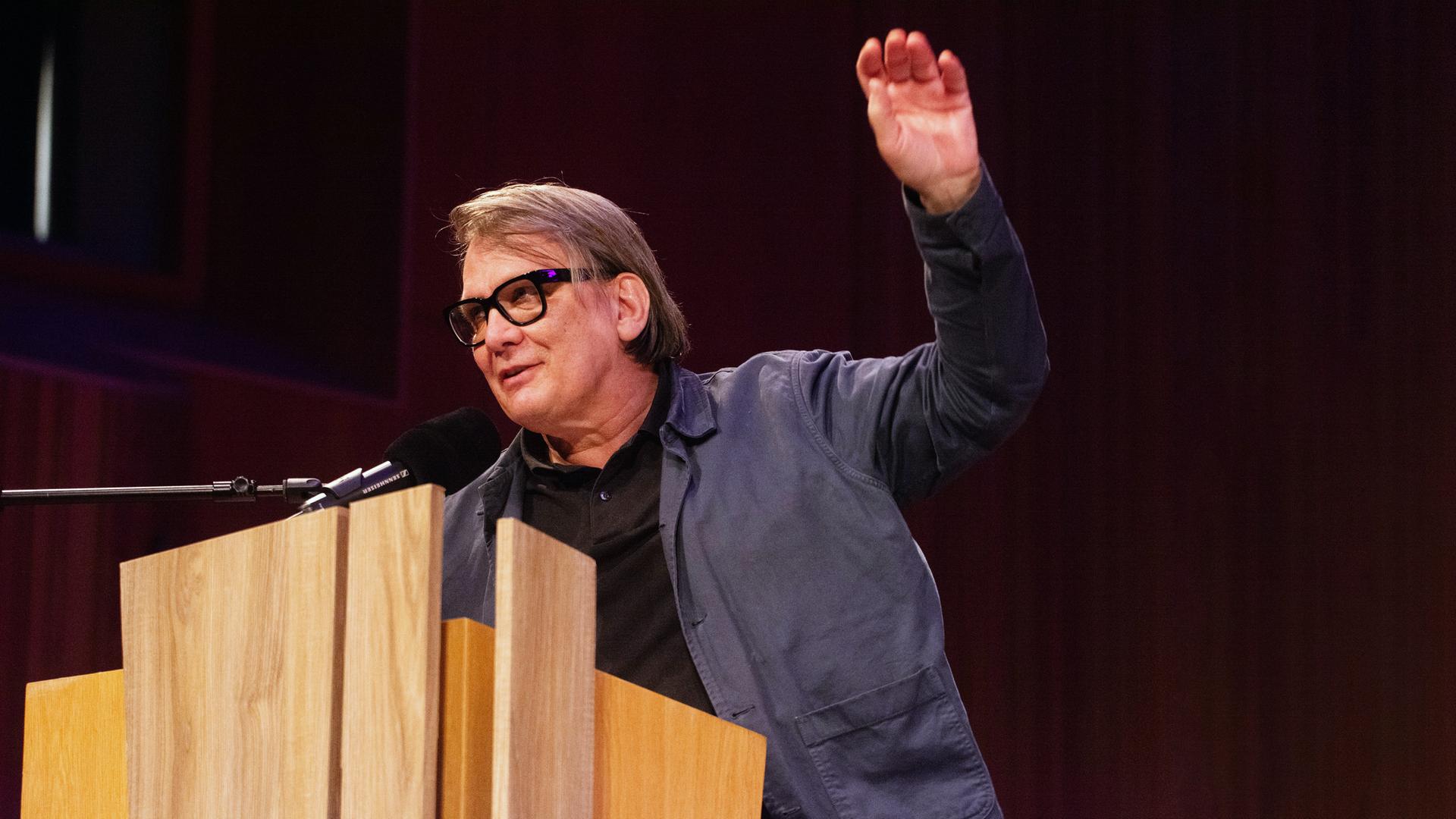 Ein Mann mit dicker schwarzer Brillenfassung spricht an einem Pult in ein Mikrofon und hebt die linke Hand.