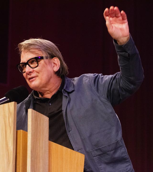 Ein Mann mit dicker schwarzer Brillenfassung spricht an einem Pult in ein Mikrofon und hebt die linke Hand.