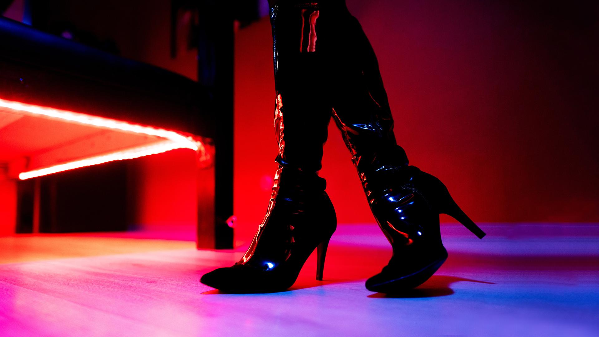 Beine in Lack-Highheel-Stiefeln stehen vor Bett in Bordell.