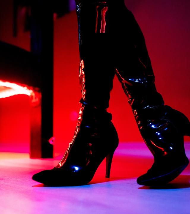 Beine in Lack-Highheel-Stiefeln stehen vor Bett in Bordell.