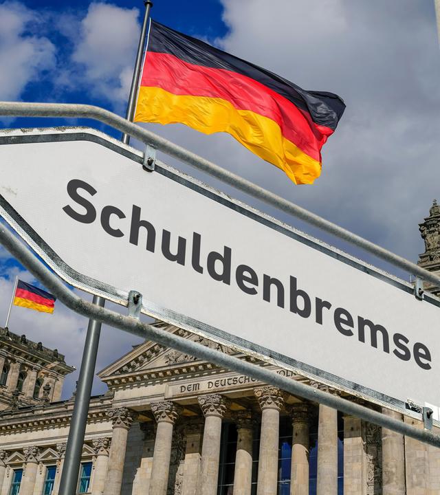 Hinter einem Straßenschild mit der Aufschrift Schuldenbremse ist der Bundestag mit wehender Deutschlandflagge zu sehen.