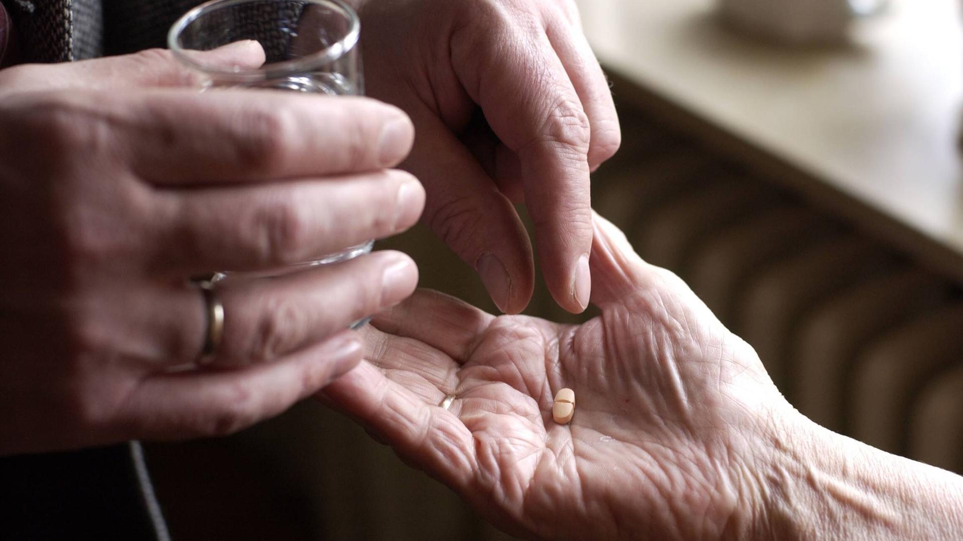 Auf dem Bild ist ein Paar Hände zu sehen. Eine Hand hält ein Wasserglas, während die andere eine Pille in die Hand einer alten Frau legt.