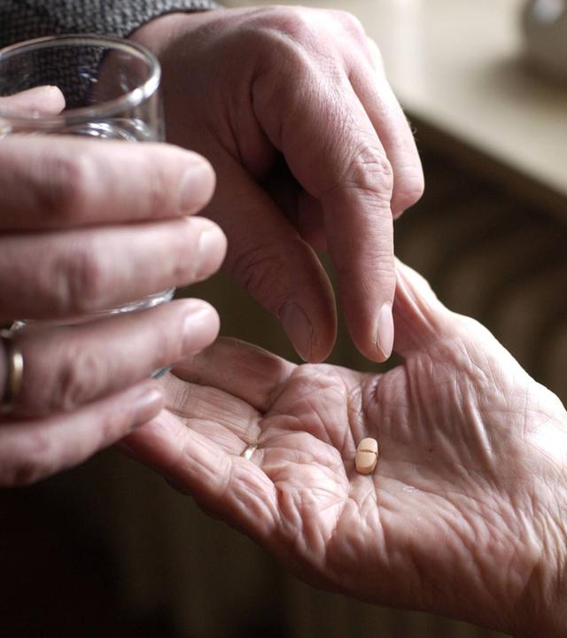 Auf dem Bild ist ein Paar Hände zu sehen. Eine Hand hält ein Wasserglas, während die andere eine Pille in die Hand einer alten Frau legt.