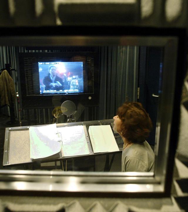 Eine Frau sitzt hinter einer Glasscheibe in einem kleinen Studio und spricht in ein Mikrofon, während sie auf einem Bildschirm einen Film schaut.