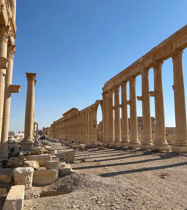antike Säulen stehen vor blauem Himmel im Wüstensand.