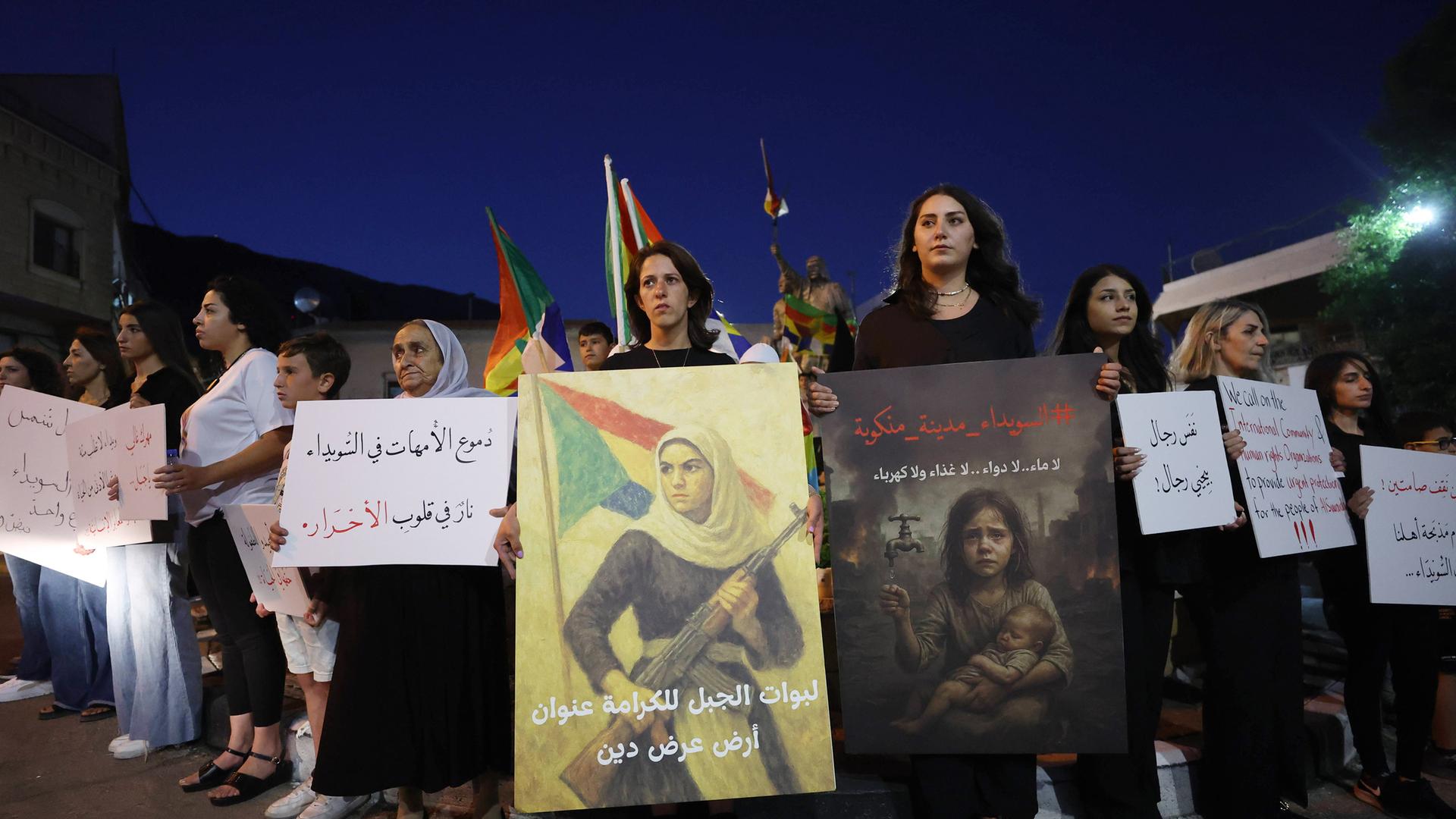 Mehrere Menschen halten Banner und Plakate bei einer Demonstration im Dunkeln.